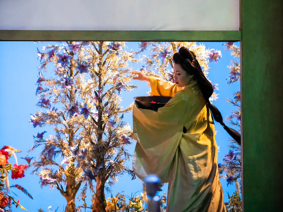 A performer wearing a kimono holds a basket in one arm and tends to tall flowers with the other on a well-lit stage with a bright blue backdrop. They are performing in The Royal Opera's production of Madama Butterfly.