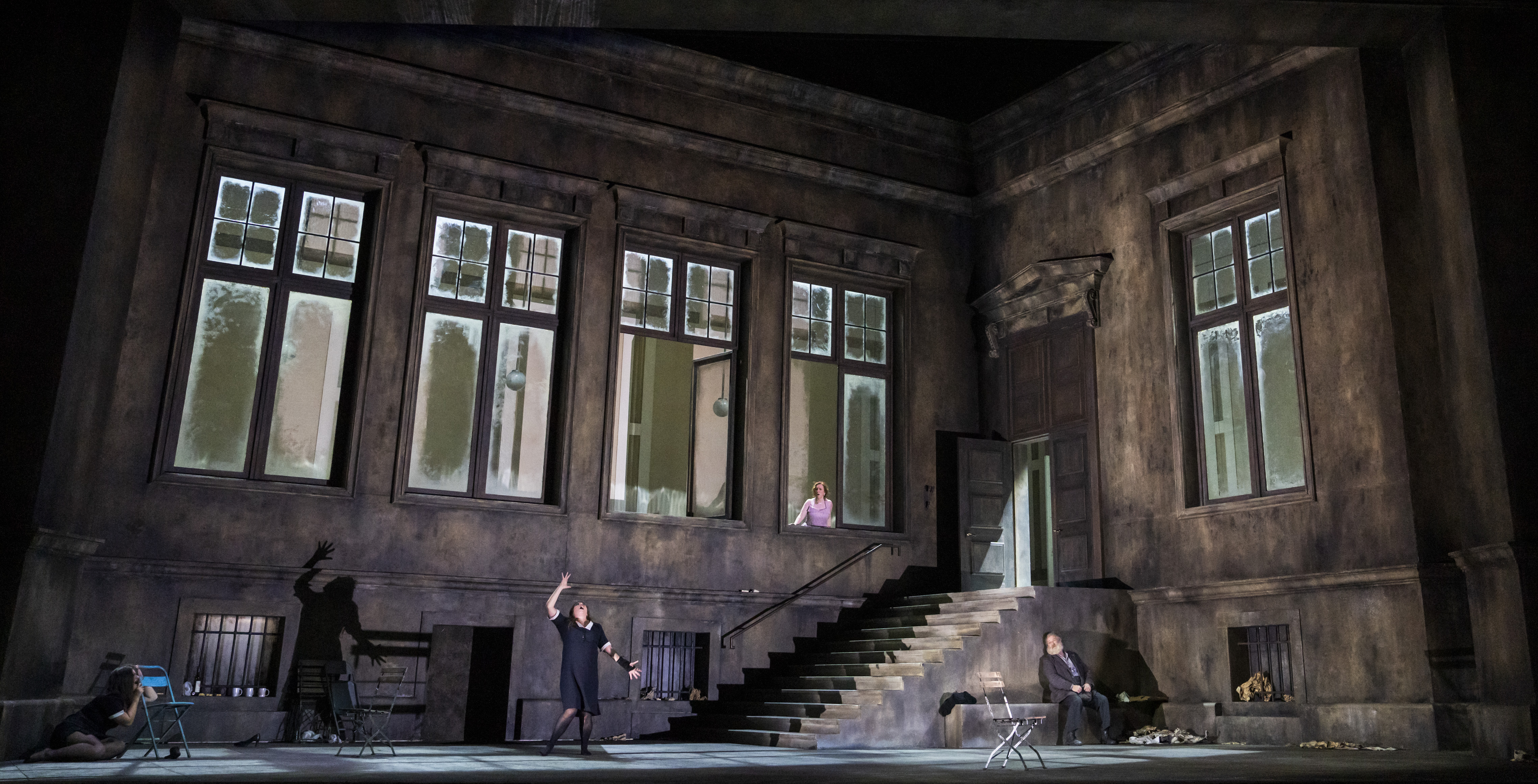 A performer wearing a black and white maids uniform stands on stage in the grounds of a large building with stairs leading up. They hold their arm upwards which makes a shadow silhouette on the wall behind. (12 of 12)