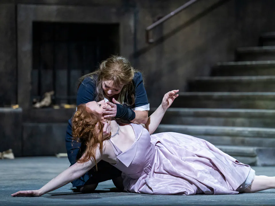 A performer wearing a black and white maids uniform holds the head up of a lifeless-looking performer wearing a purple dress.