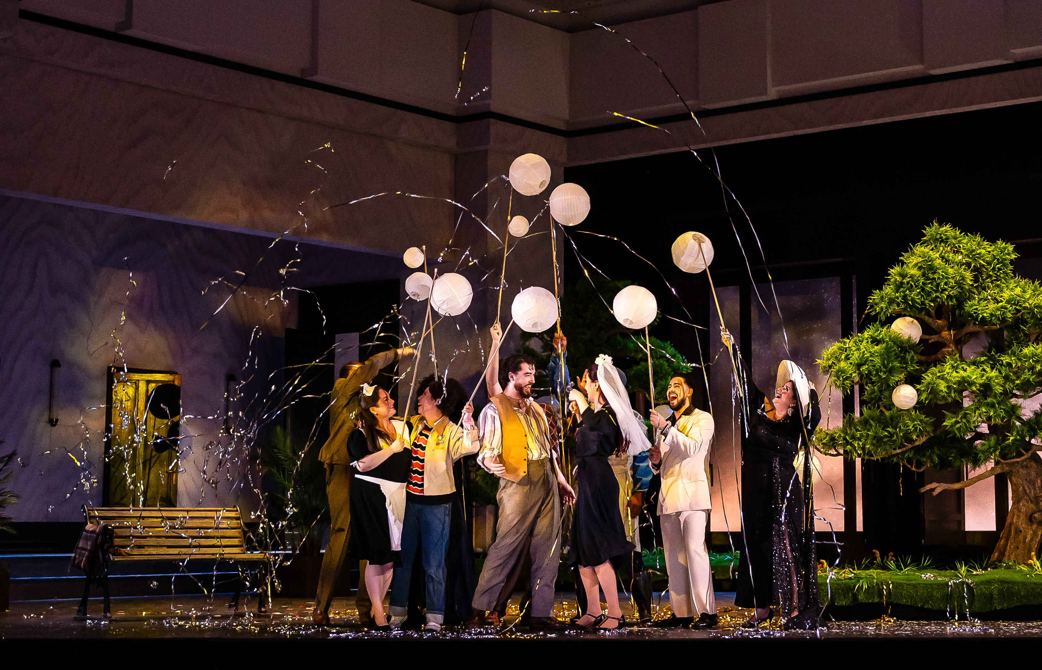 A group of performers stand on a stage set with a bench and a bonsai tree. They are in celebration using party poppers. 