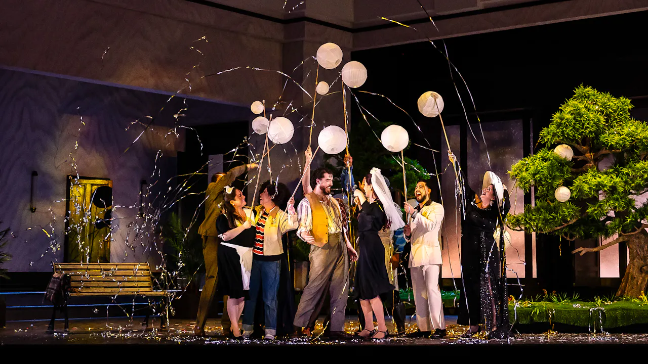 A group of performers stand on a stage set with a bench and a bonsai tree. They are in celebration using party poppers.