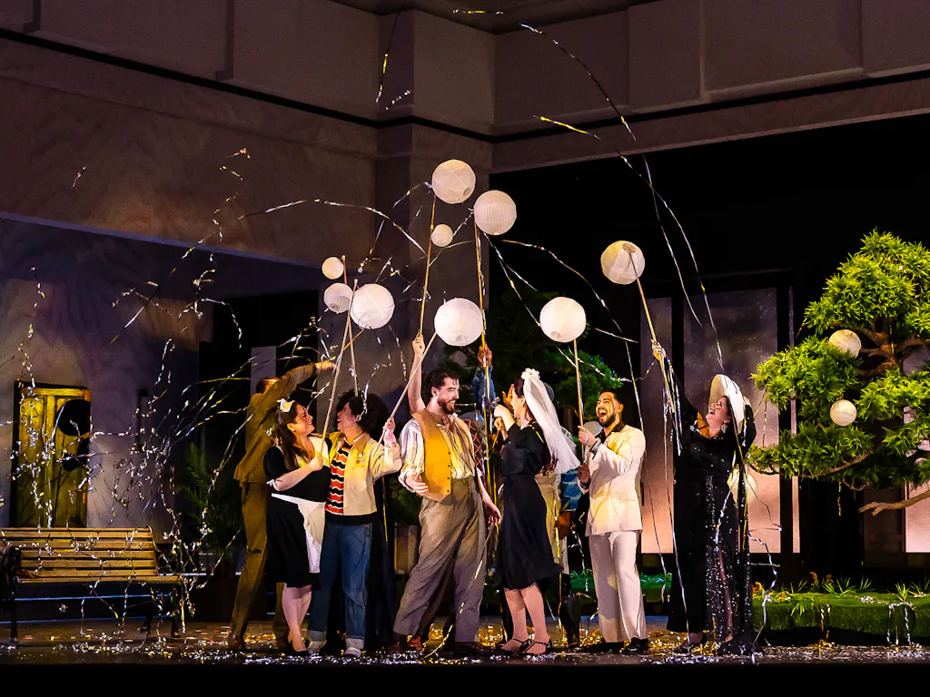 A group of performers stand on a stage set with a bench and a bonsai tree. They are in celebration using party poppers.