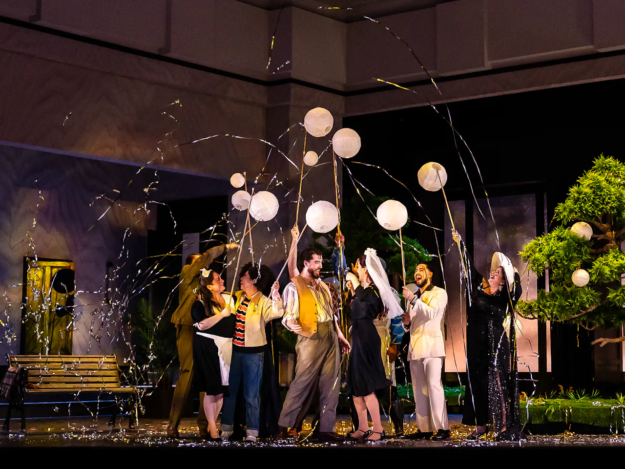 A group of performers stand on a stage set with a bench and a bonsai tree. They are in celebration using party poppers.