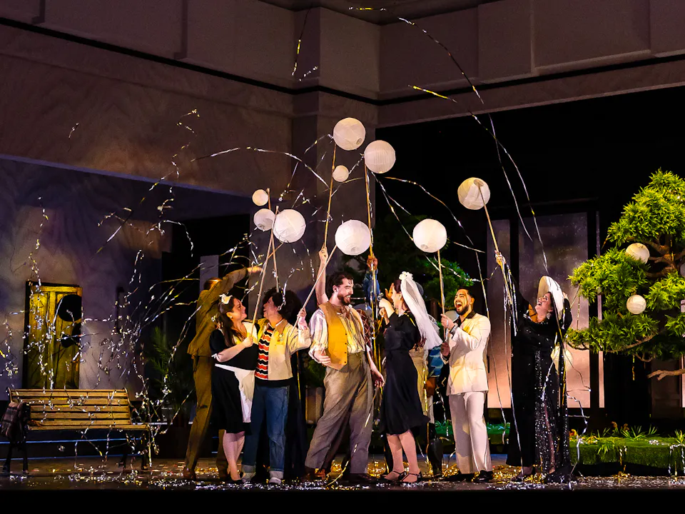 A group of performers stand on a stage set with a bench and a bonsai tree. They are in celebration using party poppers.
