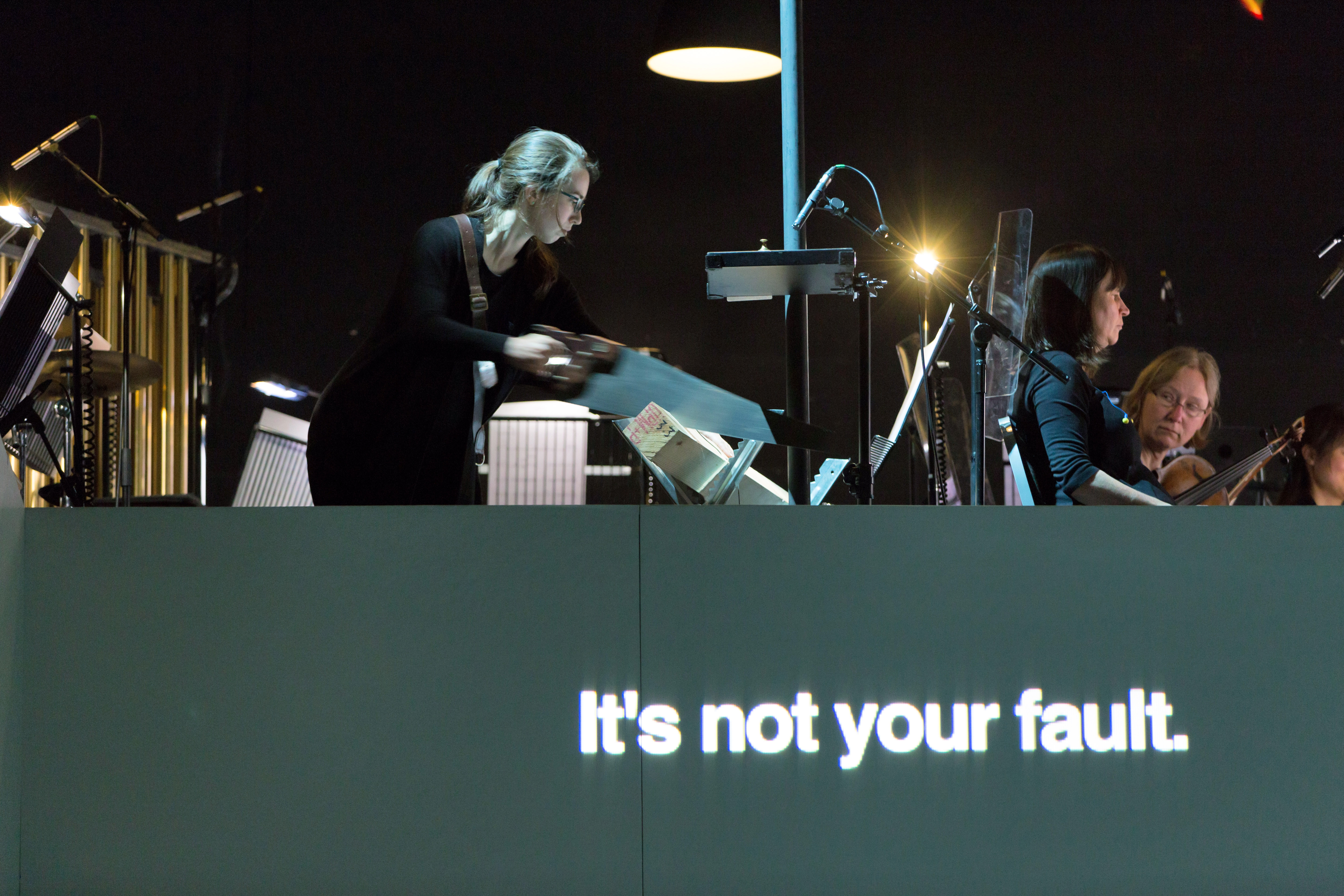 A view from a dark orchestra put as a conductors motions to musicians. The image is artistically blurred and altered in parts. The words 'It's not your fault." are written in white on grey at the bottom. (2 of 4)