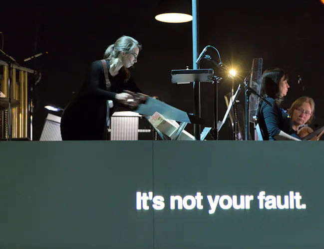 A view from a dark orchestra put as a conductors motions to musicians. The image is artistically blurred and altered in parts. The words 'It's not your fault." are written in white on grey at the bottom. (2 of 4)