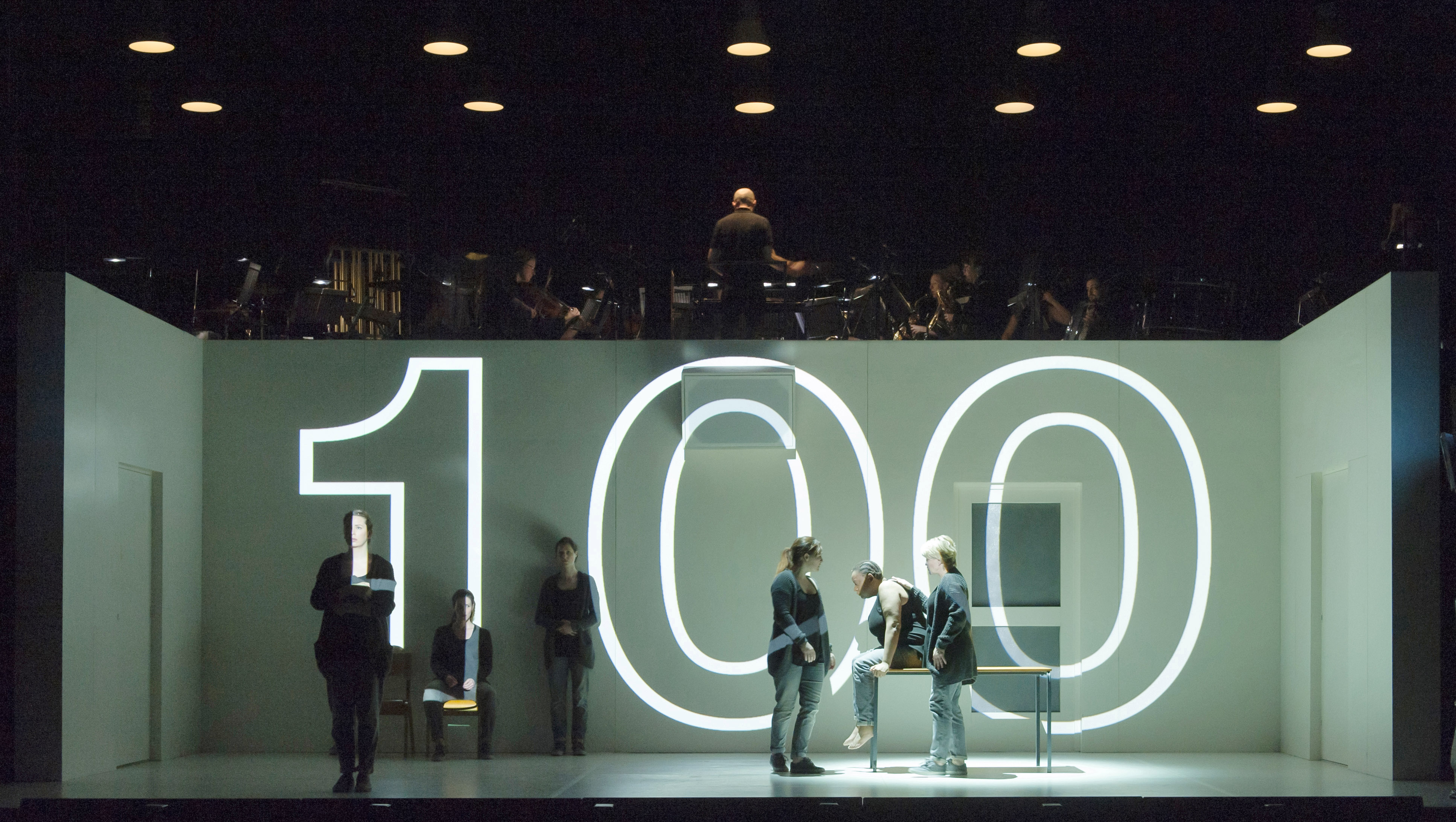 Several performers move around a stage within a white dimly-lit room with doors and windows. Above the room are musicians from an orchestra. The number 100 is projected in bright white lights directly onto the room space. (3 of 12)