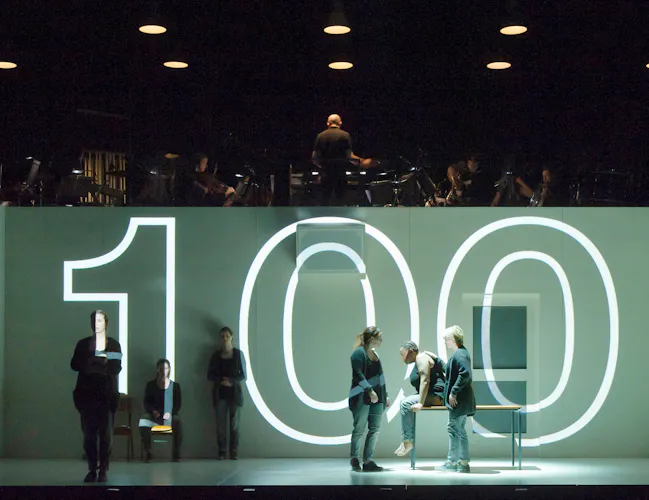 Several performers move around a stage within a white dimly-lit room with doors and windows. Above the room are musicians from an orchestra. The number 100 is projected in bright white lights directly onto the room space. (3 of 12)