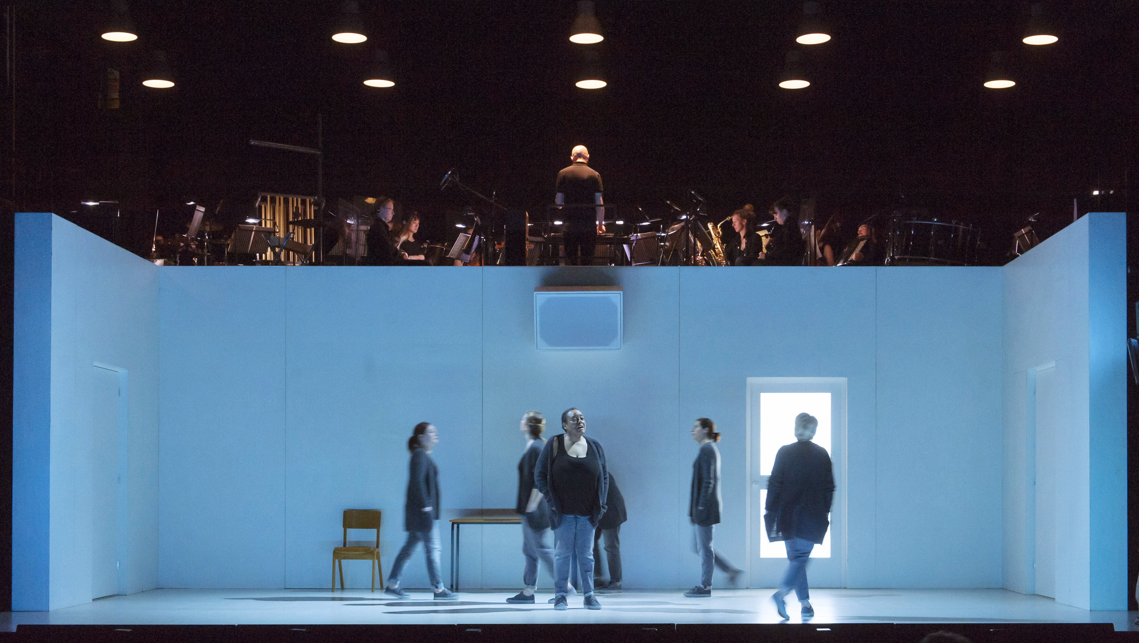 Several performers move around on stage within a blue dimly lit room with doors and windows. Above the room are musicians from an orchestra.