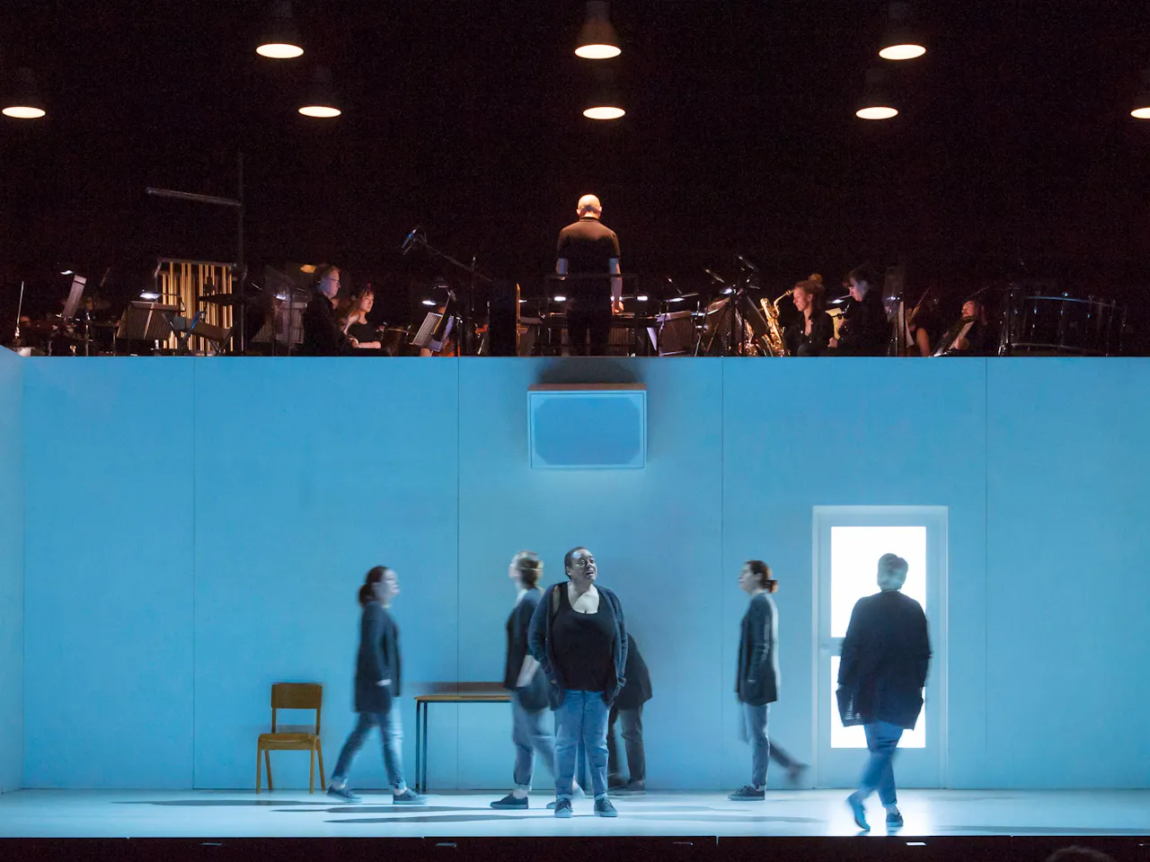 Several performers move around on stage within a blue dimly lit room with doors and windows. Above the room are musicians from an orchestra.