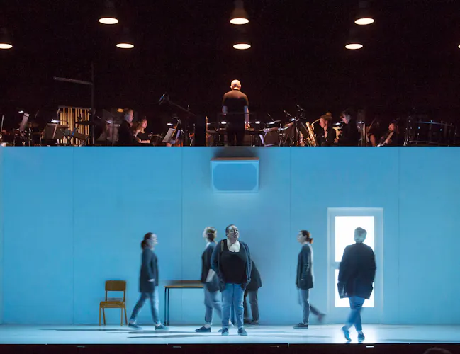 Several performers move around on stage within a blue dimly lit room with doors and windows. Above the room are musicians from an orchestra. (3 of 4)