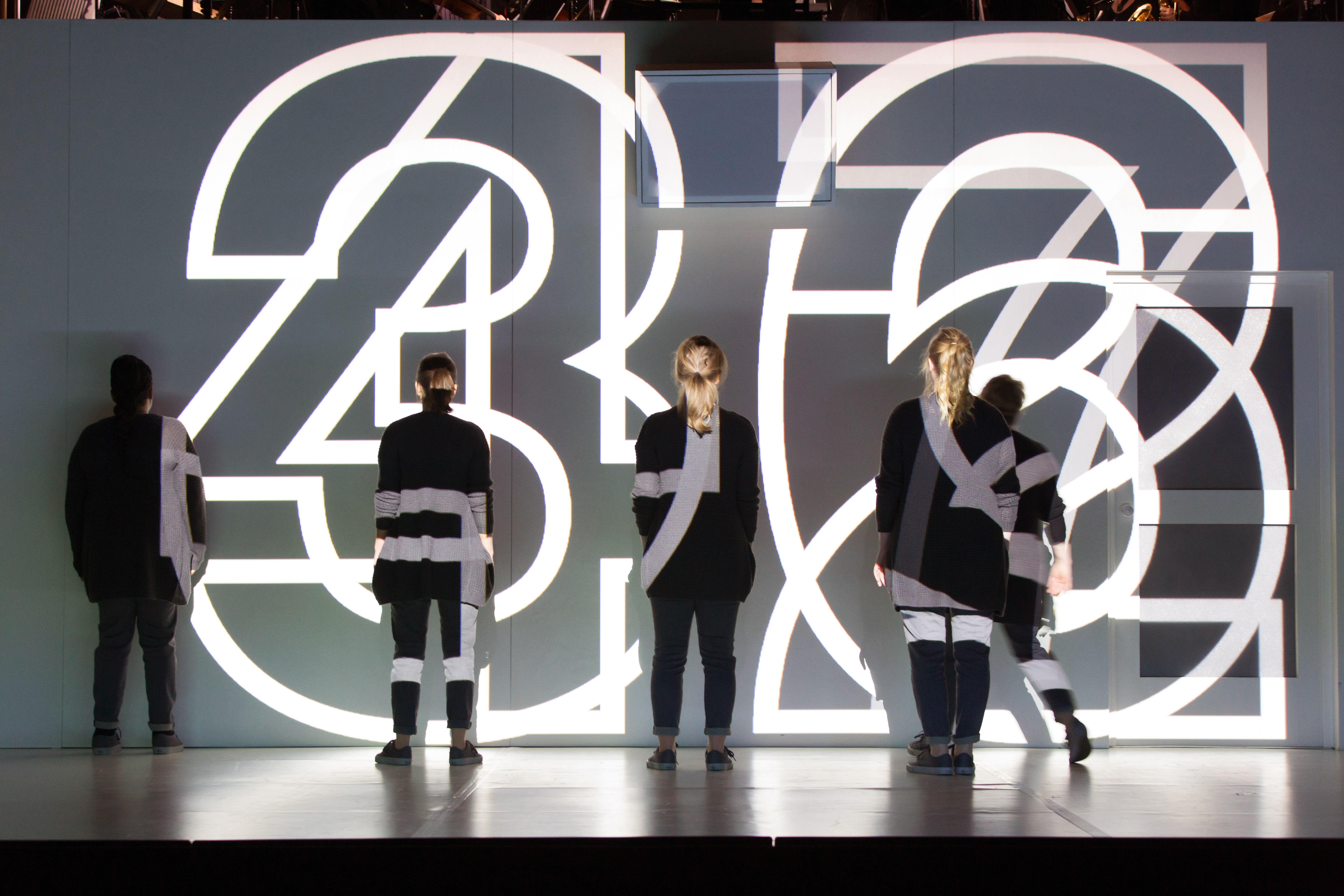 Four performers wearing black stand on stage with their backs to the audience. Large white numbers are brightly lit and projected directly onto the stage and performers. The numbers are layered on top of each other making it hard to read them properly. 