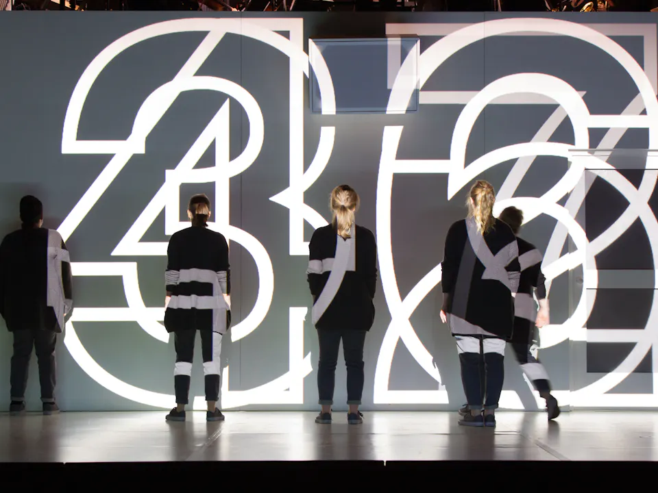 Four performers wearing black stand on stage with their backs to the audience. Large white numbers are brightly lit and projected directly onto the stage and performers. The numbers are layered on top of each other making it hard to read them properly.