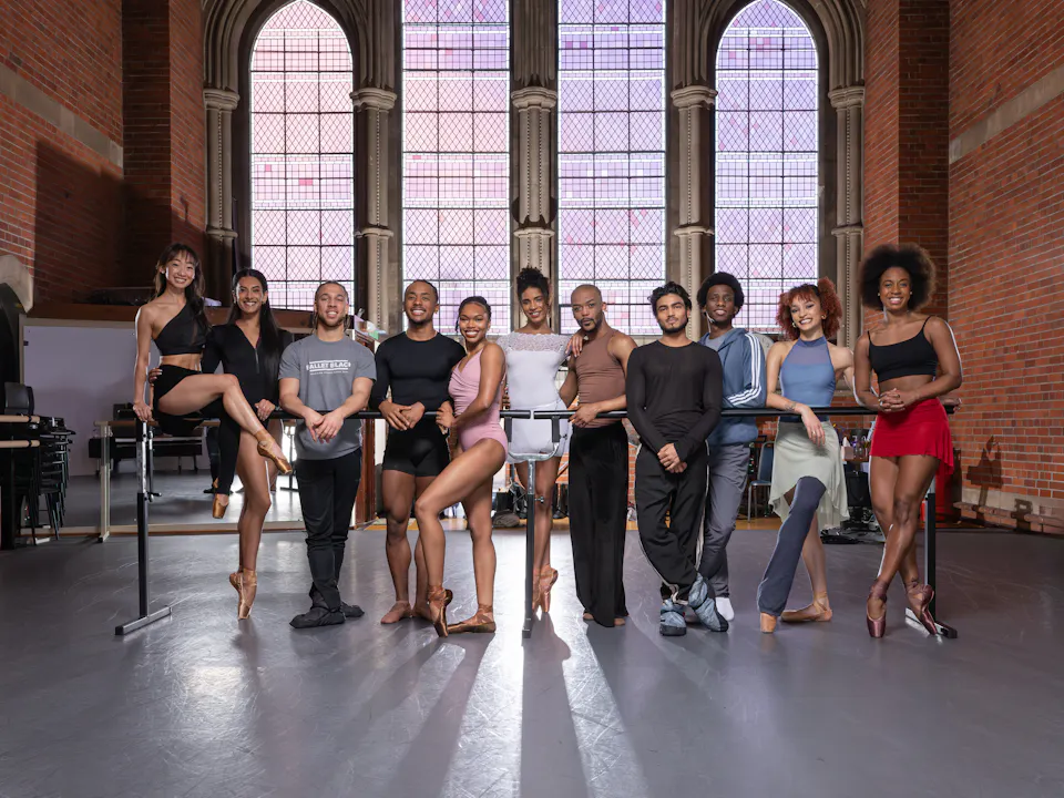 Eleven ballet dancers who are part of Ballet Black pose together in an ornate building in front of stained glass windows.