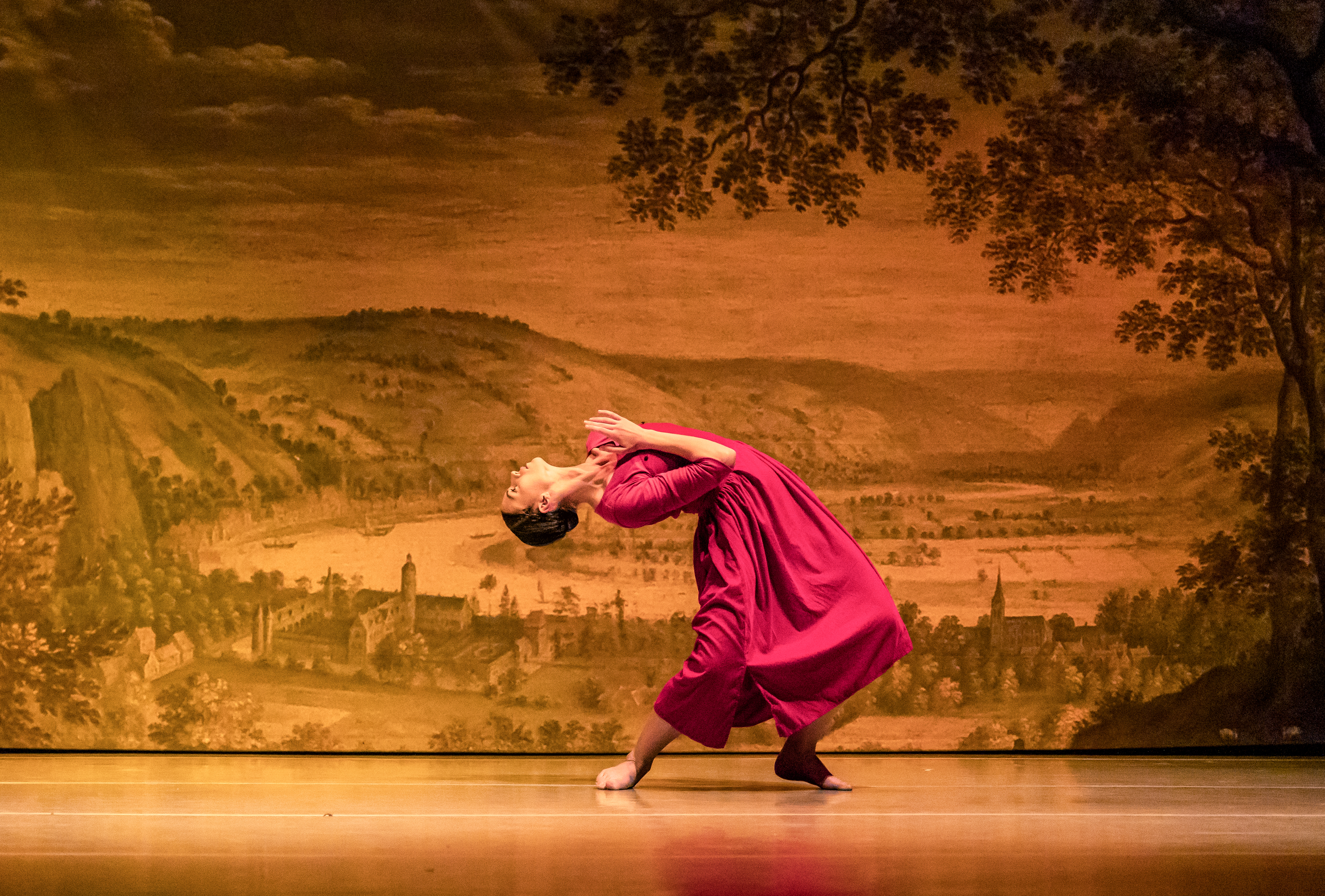 A ballet dancers performs on stage in a red dress in front of a backdrop of an old style painting of a town within a rural scene. 