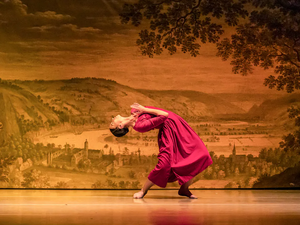 A ballet dancers performs on stage in a red dress in front of a backdrop of an old style painting of a town within a rural scene.