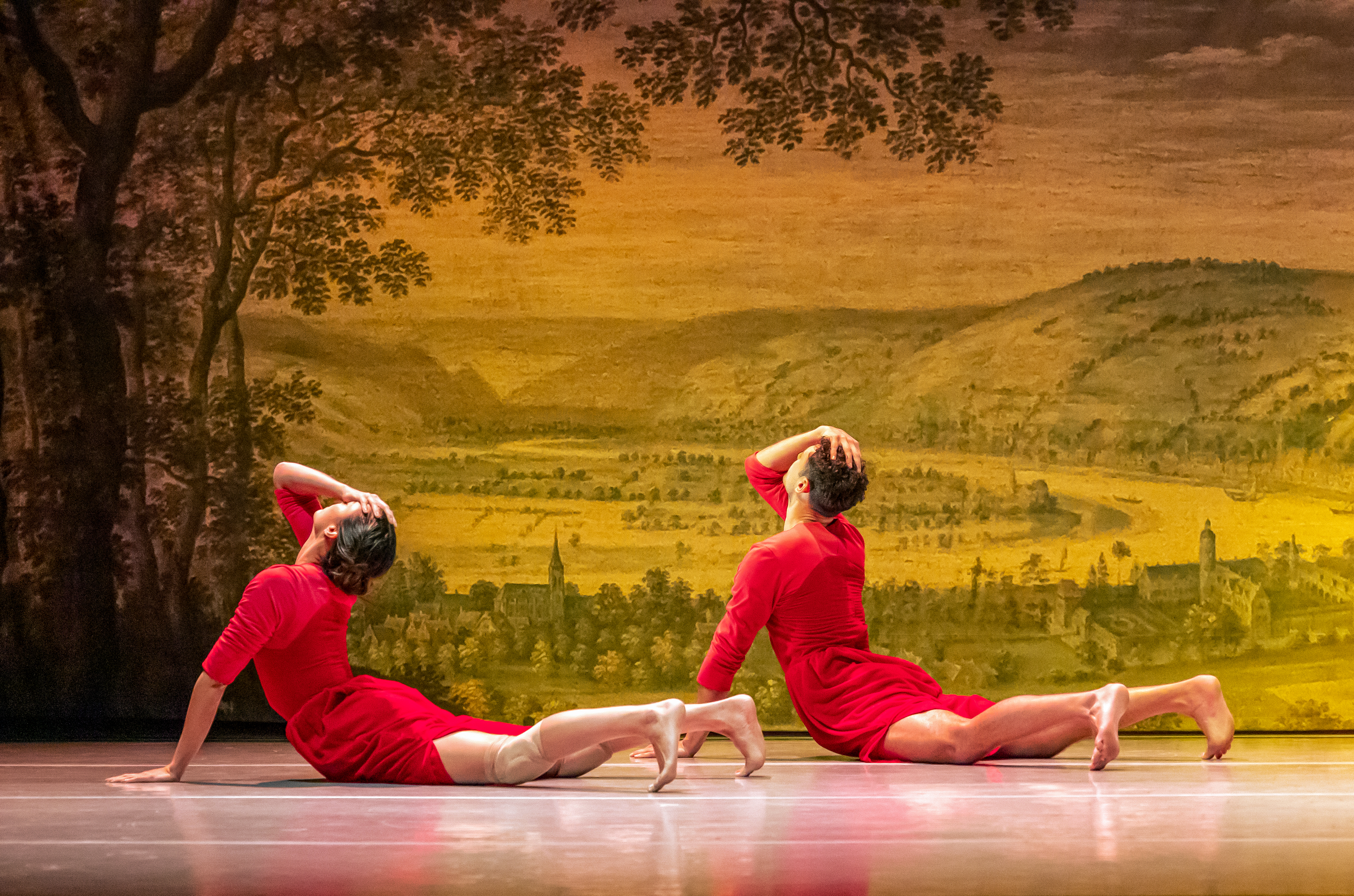 Two ballet dancers lie on their front and hold their bodies upwards wearing red in front of a backdrop of an old style painting of a town within a rural scene. (1 of 5)