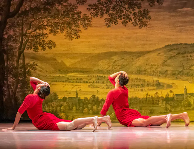 Two ballet dancers lie on their front and hold their bodies upwards wearing red in front of a backdrop of an old style painting of a town within a rural scene. (1 of 5)