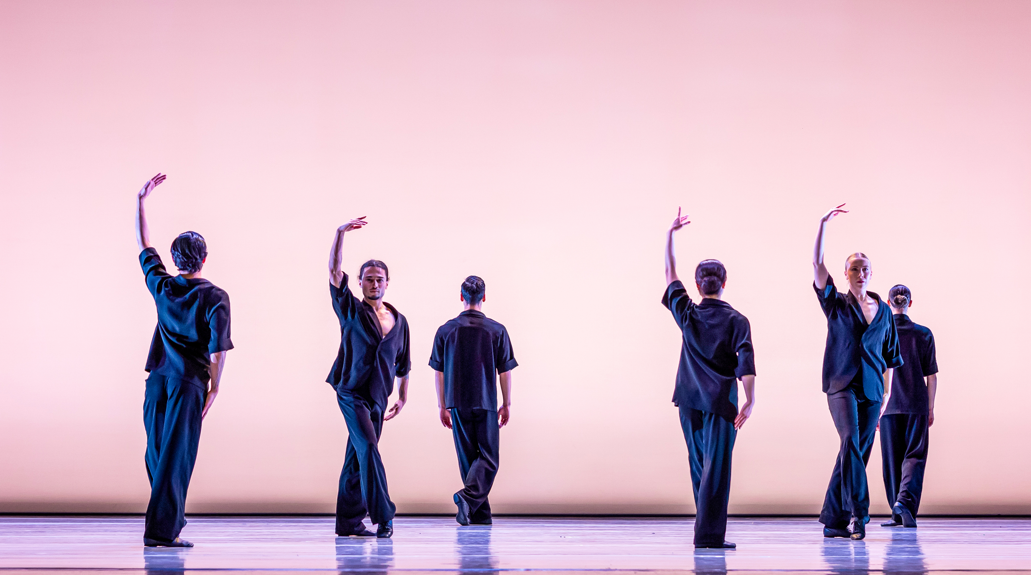 Six ballet dancers perform on a well-lit stage in front of a pink backdrop. They are wearing blue trousers and shirt. Some hold an arm upwards. (2 of 5)