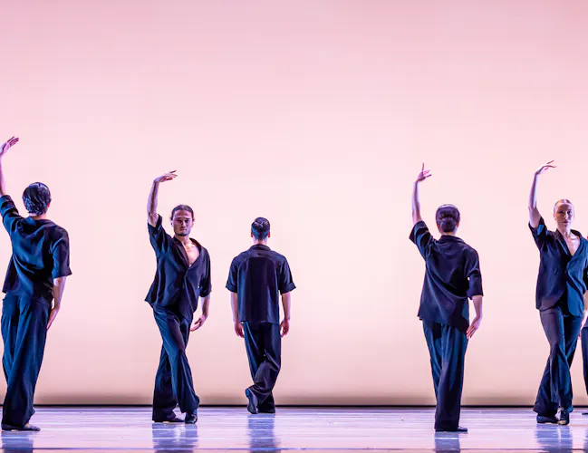 Six ballet dancers perform on a well-lit stage in front of a pink backdrop. They are wearing blue trousers and shirt. Some hold an arm upwards. (2 of 5)