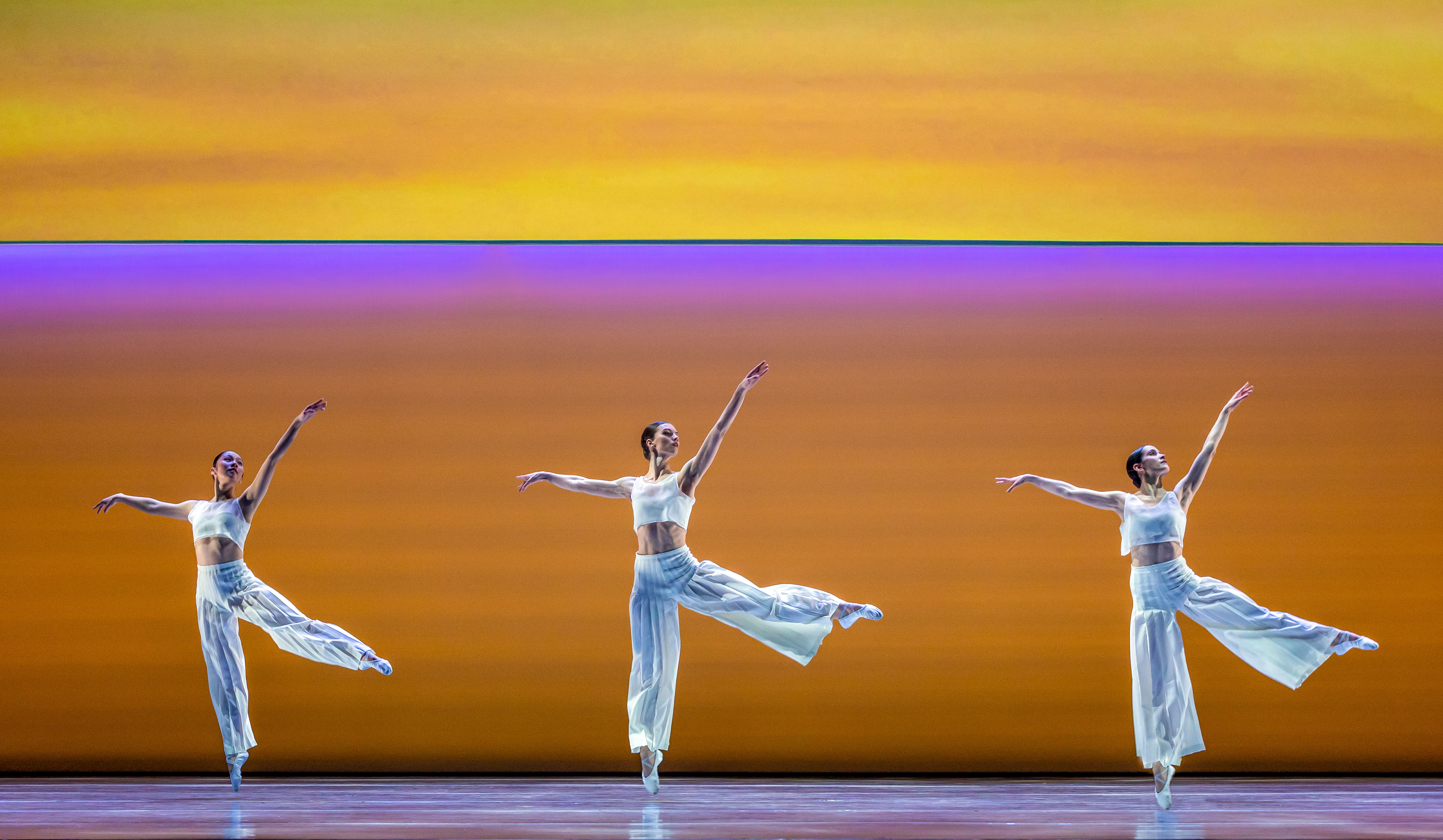 Three ballet dancers perform on stage in front of a gradient colour background which has a pink stripe running through the middle. The dancers wear all white and stand en pointe with their arms raised upwards. (3 of 5)