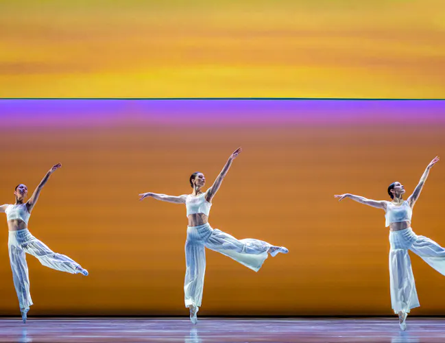 Three ballet dancers perform on stage in front of a gradient colour background which has a pink stripe running through the middle. The dancers wear all white and stand en pointe with their arms raised upwards. (3 of 5)