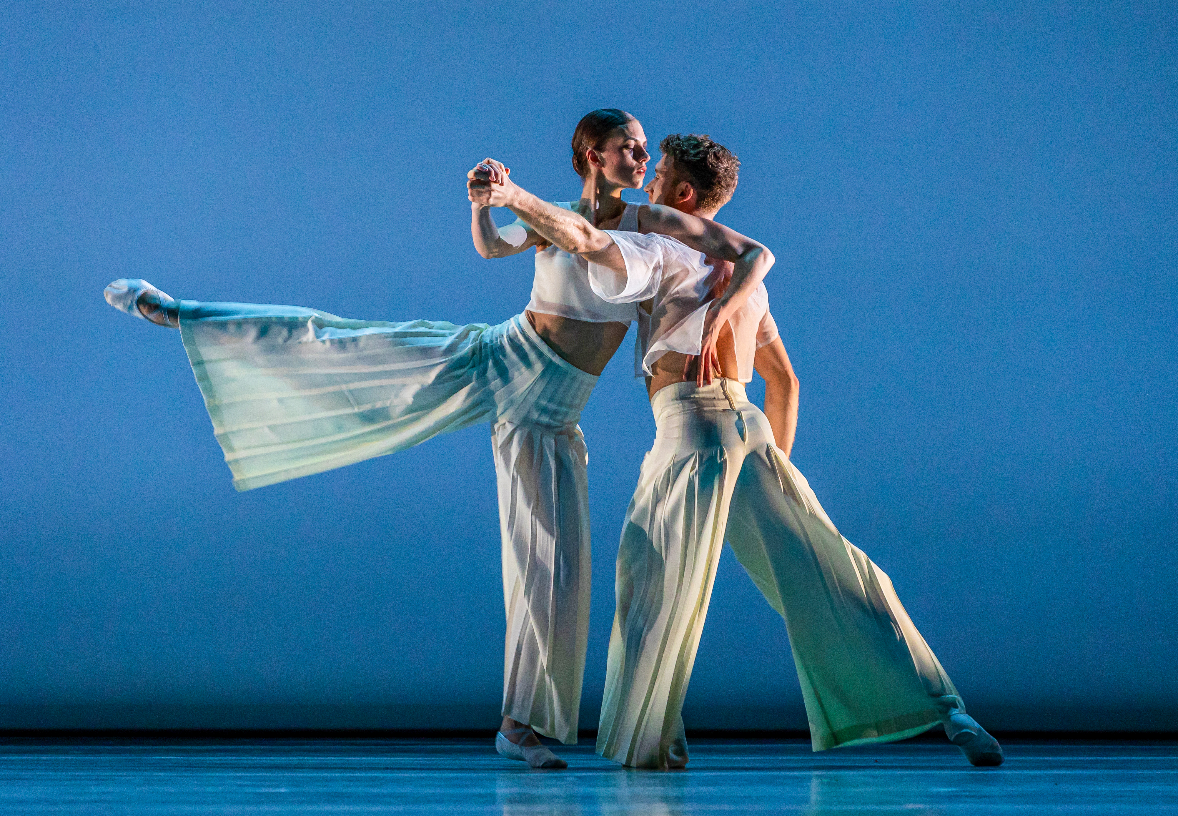 Two ballet dancers perform together in an embrace on a well-lit stage with a blue backdrop. (4 of 5)