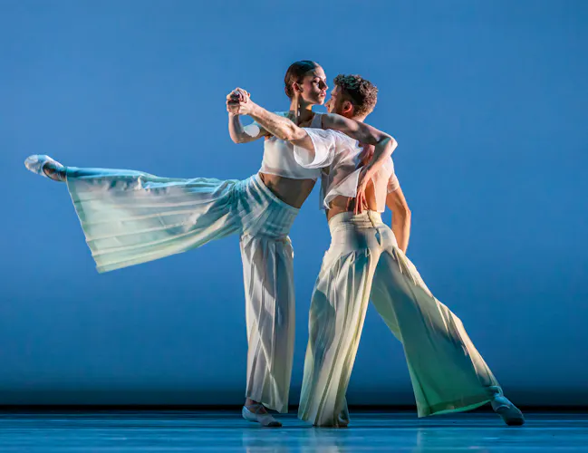 Two ballet dancers perform together in an embrace on a well-lit stage with a blue backdrop. (4 of 5)