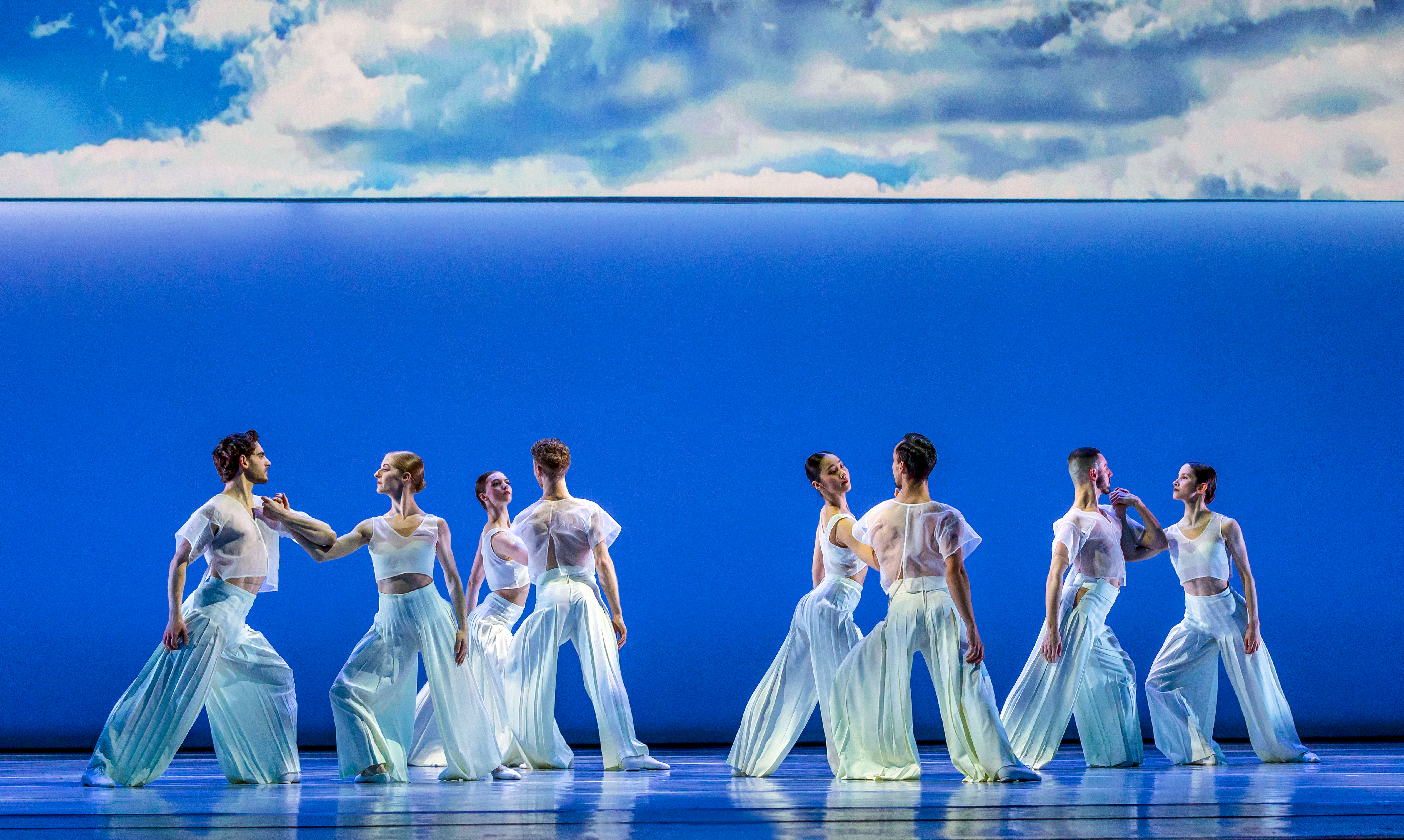 Several ballet dancers perform together on a well-lit stage with a blue backdrop where half of the backdrop is made up of a painting of the sky. (5 of 5)