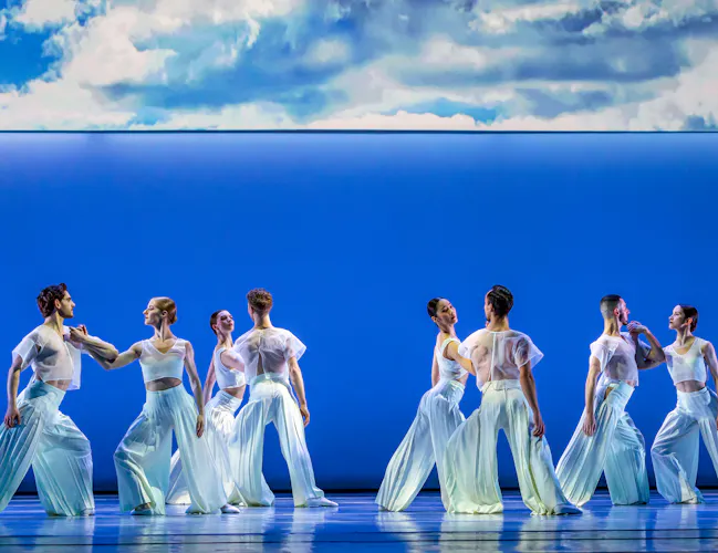 Several ballet dancers perform together on a well-lit stage with a blue backdrop where half of the backdrop is made up of a painting of the sky. (5 of 5)