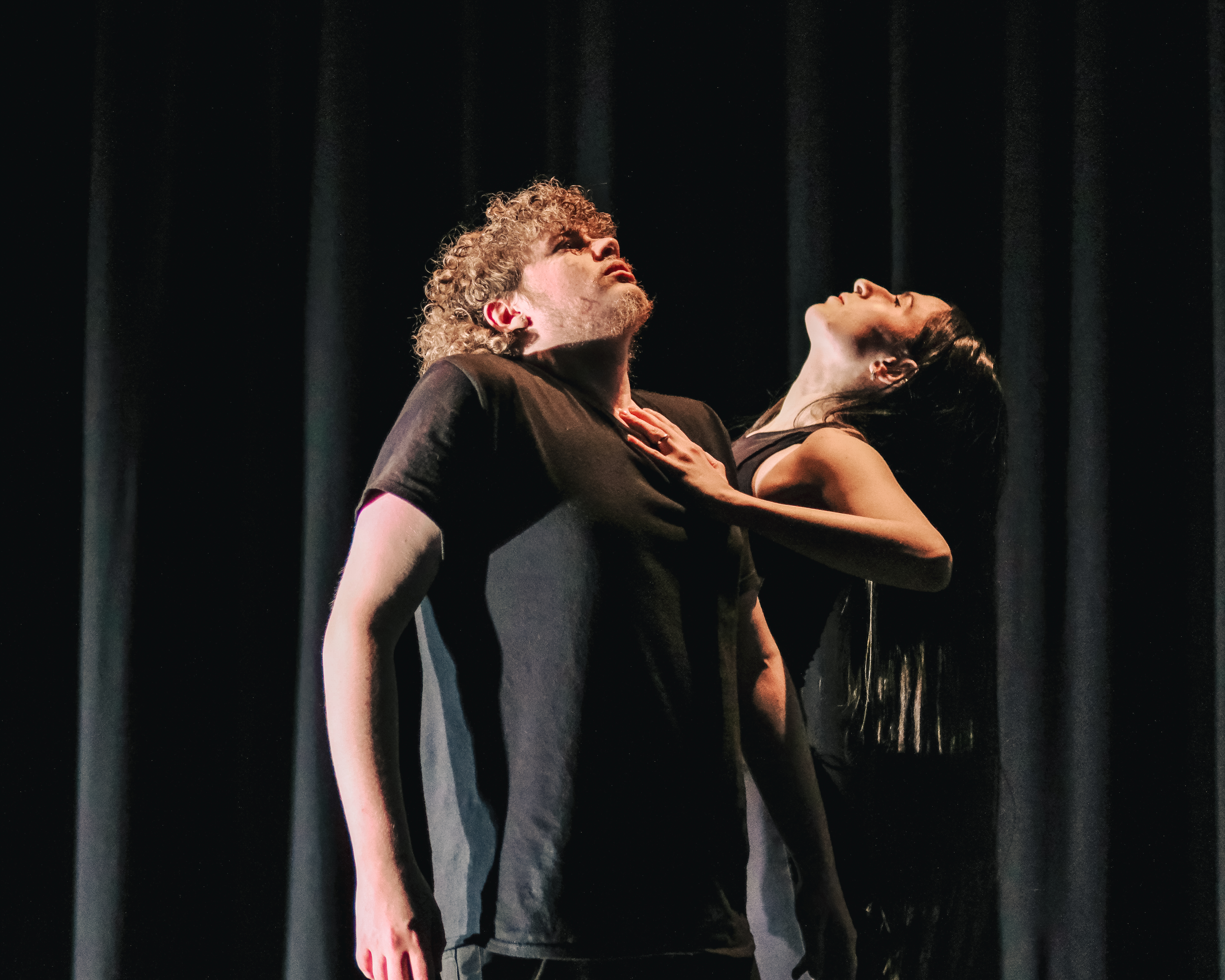 Two ballet dancers wearing black stand on stage in front of a black curtain.
