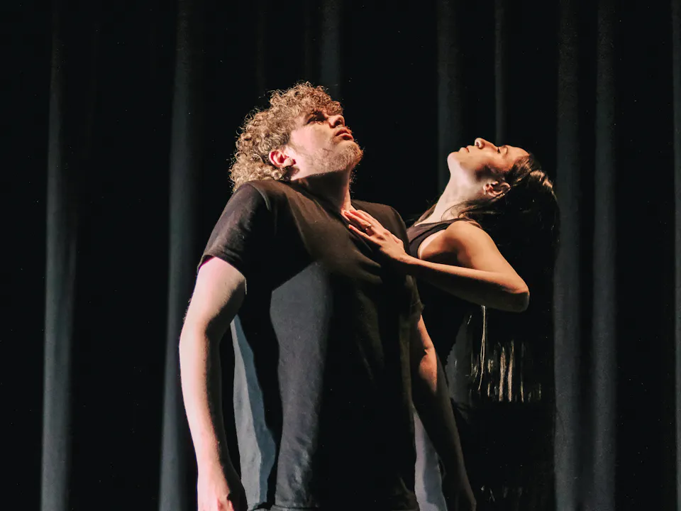 Two ballet dancers wearing black stand on stage in front of a black curtain.