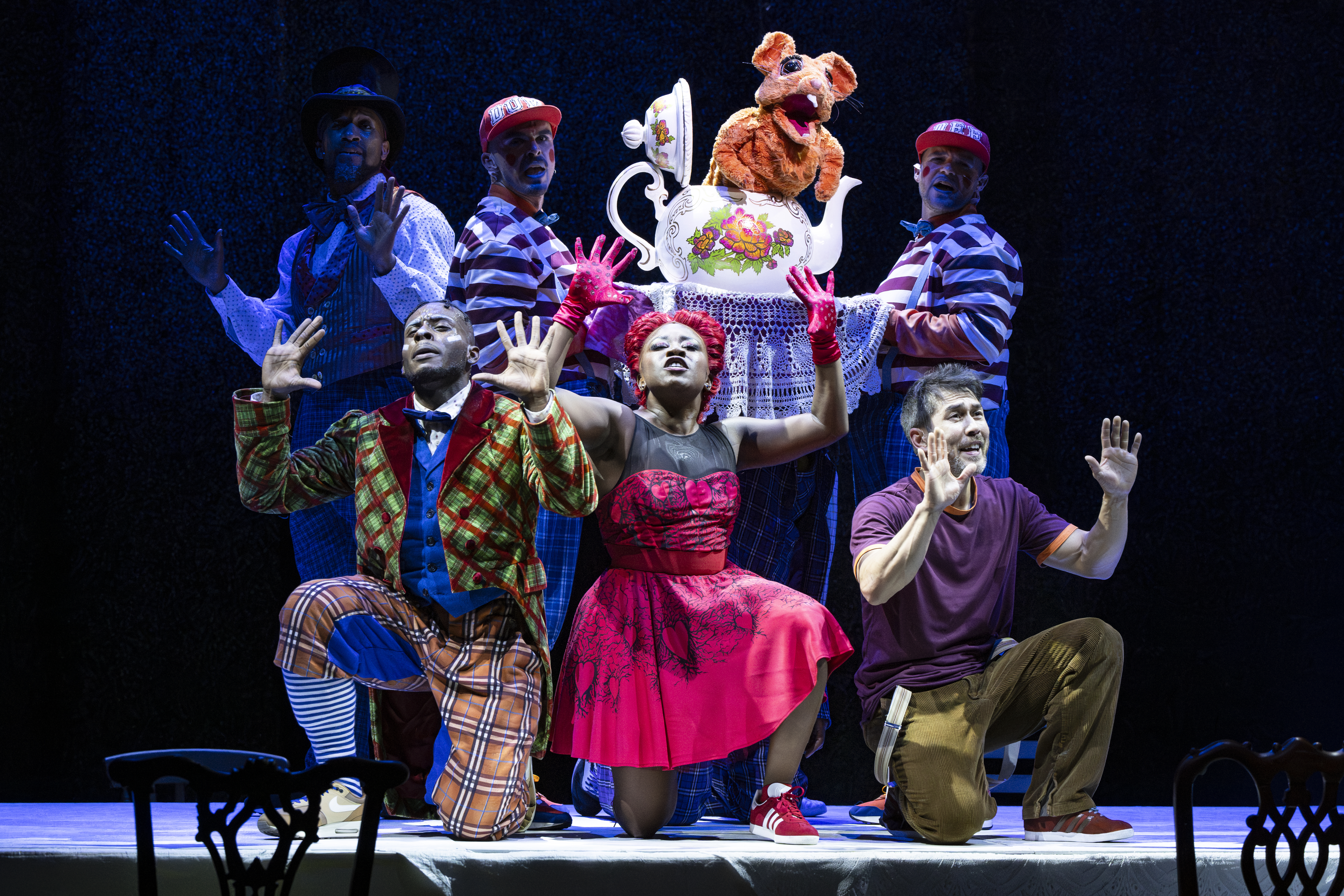 Several performers wearing bright colour costumes stand together on a dark stage. Two of the performers are wearing matching striped tops and caps and are holding up a large tea pot with a puppet of a mouse emerging from within. (1 of 4)