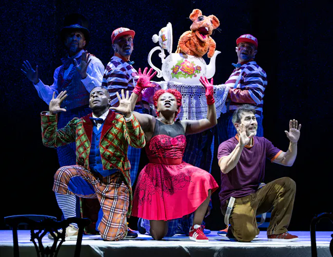 Several performers wearing bright colour costumes stand together on a dark stage. Two of the performers are wearing matching striped tops and caps and are holding up a large tea pot with a puppet of a mouse emerging from within. (1 of 4)