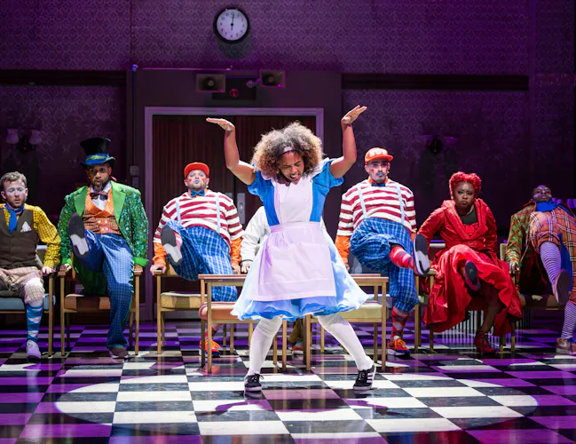 A performer wearing a blue dress with a white apron, white socks and trainers dances on stage on a checkerboard floor. Behind her, several performers wearing bright coloured costumes are dancing on chairs. (6 of 10)