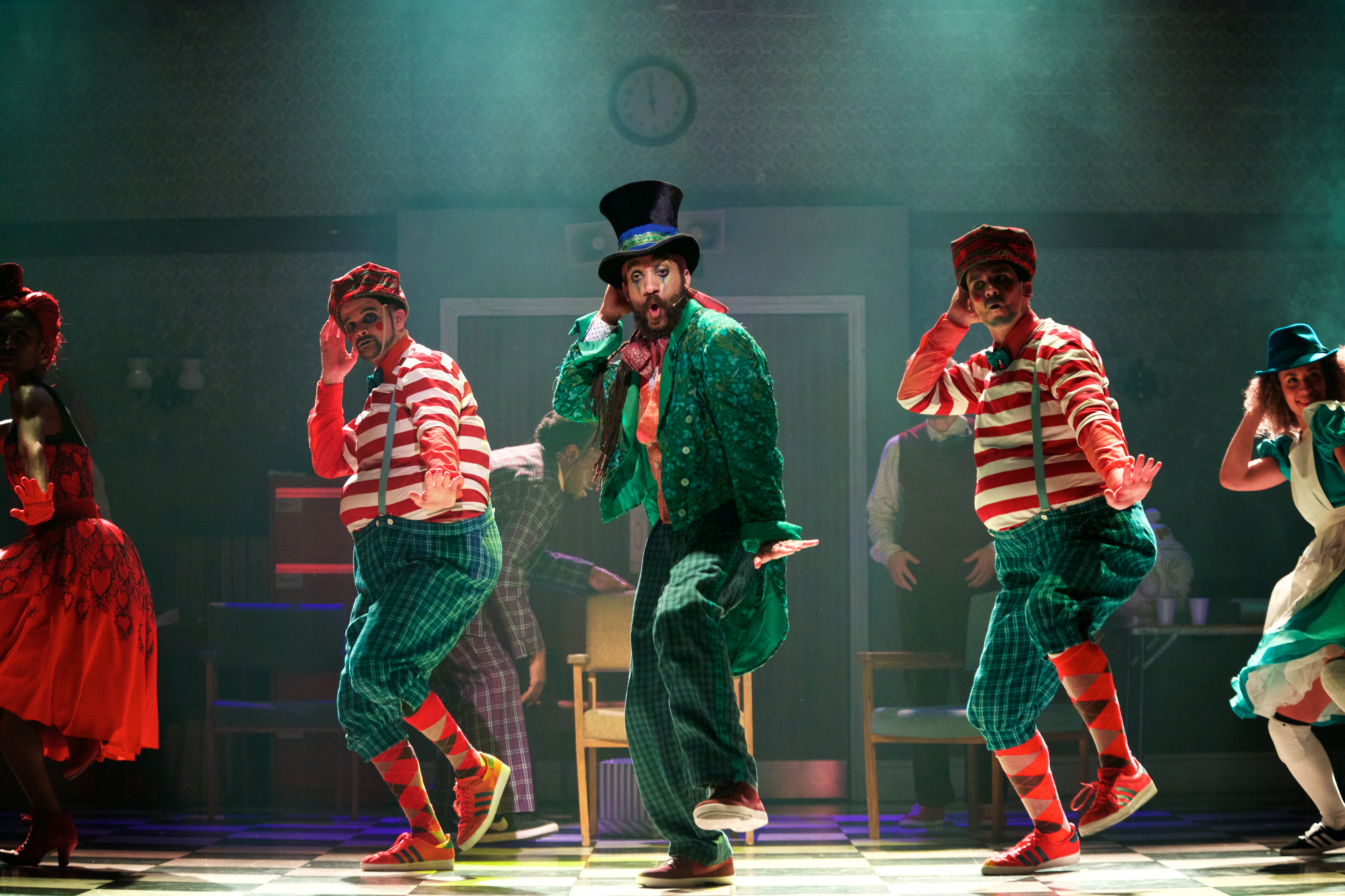 A performer wearing a green suit, bright colour top and a top hat dances on stage. They are flanked by two performers wearing matching costumes of a red striped tshirt and patterned socks. (3 of 4)