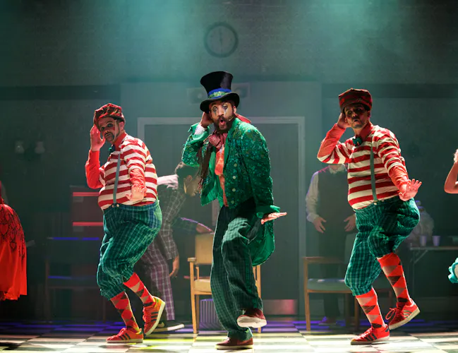A performer wearing a green suit, bright colour top and a top hat dances on stage. They are flanked by two performers wearing matching costumes of a red striped tshirt and patterned socks. (3 of 4)