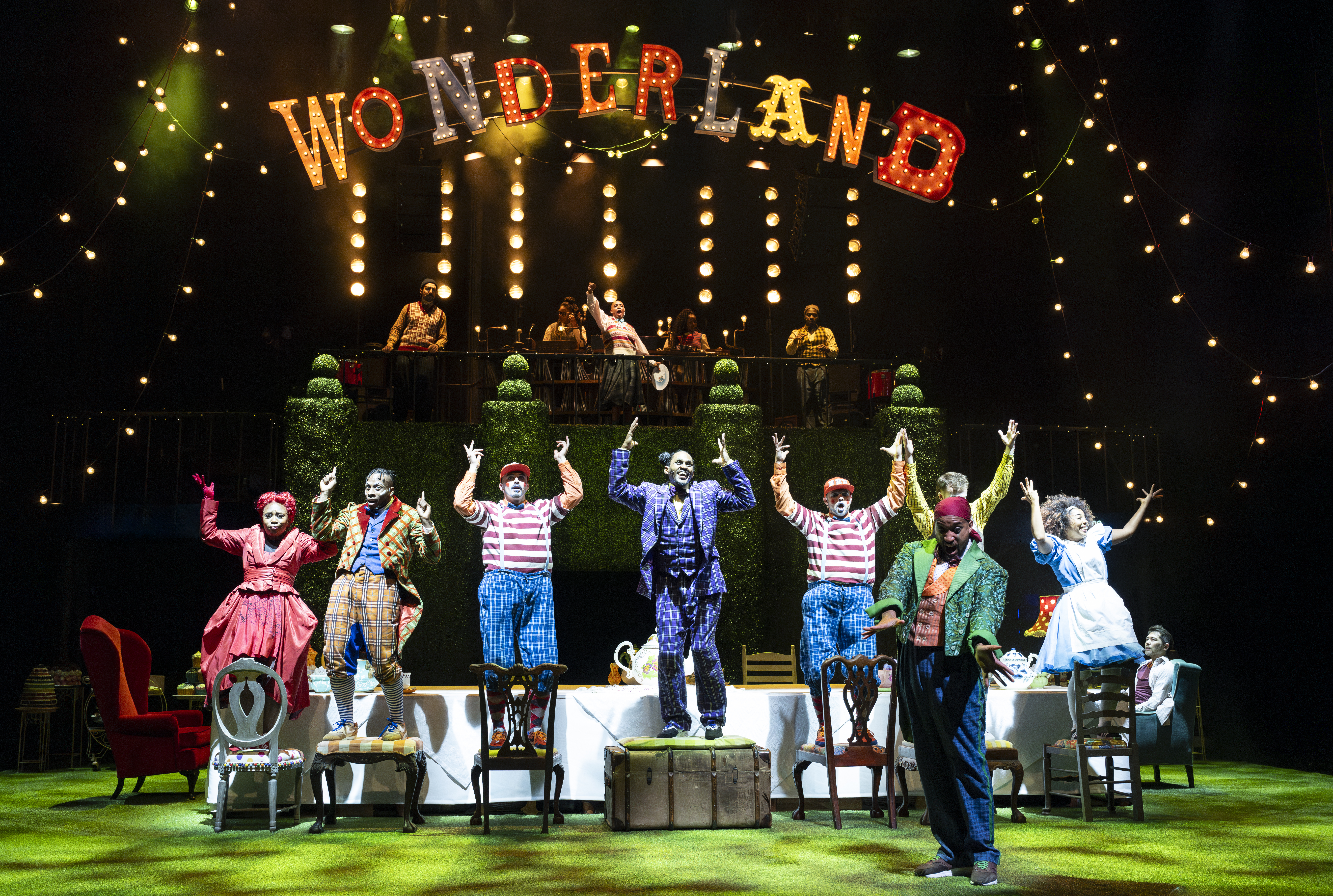 Several performers wearing bright coloured costumes stand on chairs around a long dining table. They all hold their arms in the air. Above them on a raised platform lined with shaped topiary plants is a group of musicians. The word "Wonderland" is brightly lit and raised above. 