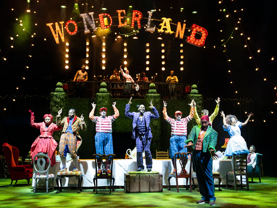 Several performers wearing bright coloured costumes stand on chairs around a long dining table. They all hold their arms in the air. Above them on a raised platform lined with shaped topiary plants is a group of musicians. The word "Wonderland" is brightly lit and raised above.