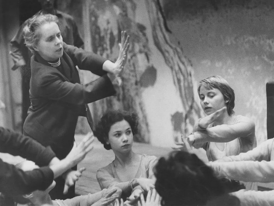 A group of women sit around holding their arms upwards placing one hand in front of the other. The image is black and white and artistic.
