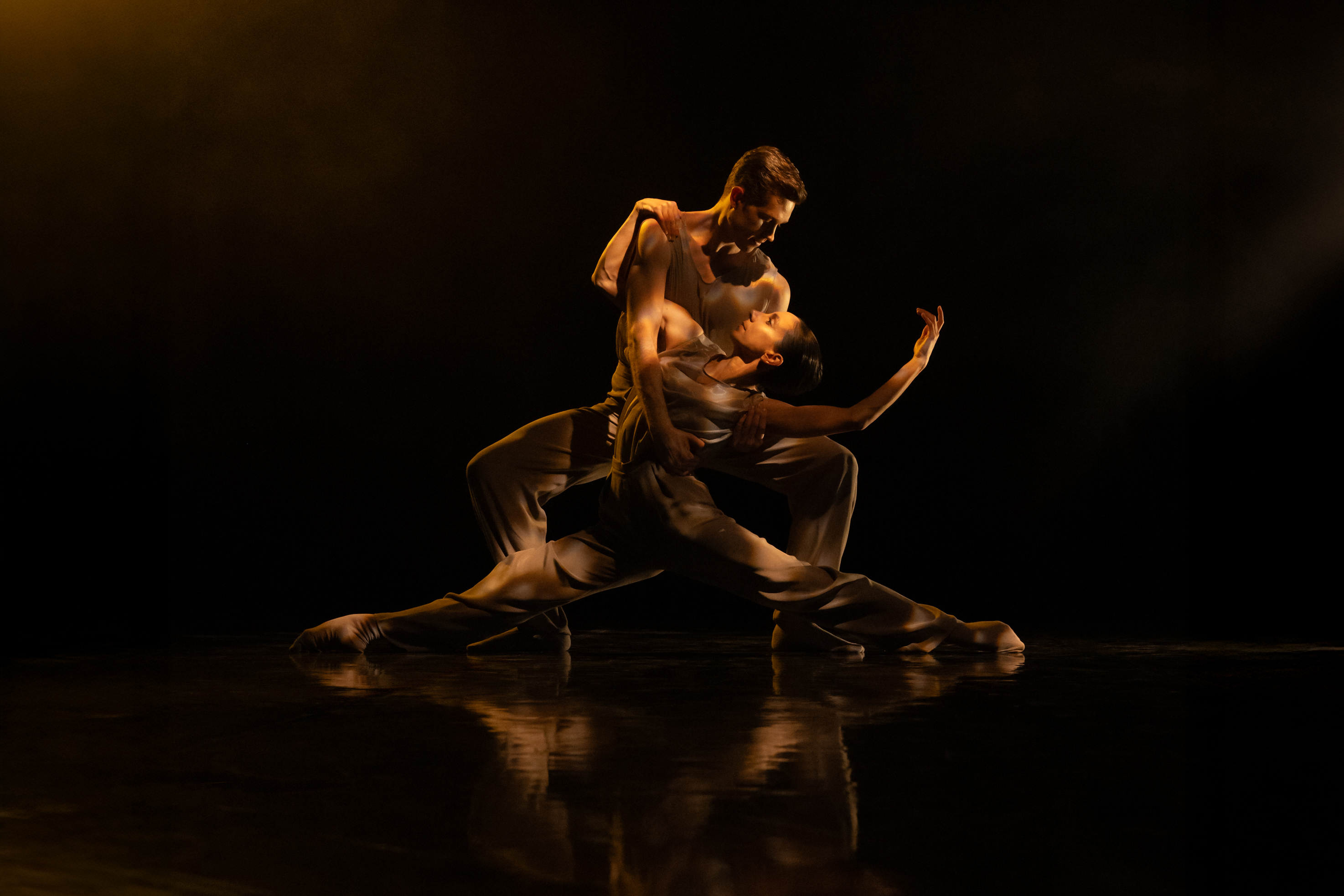 Two ballet dancers embrace passionately on a dark stage. One dancer draped themselves over the other. 