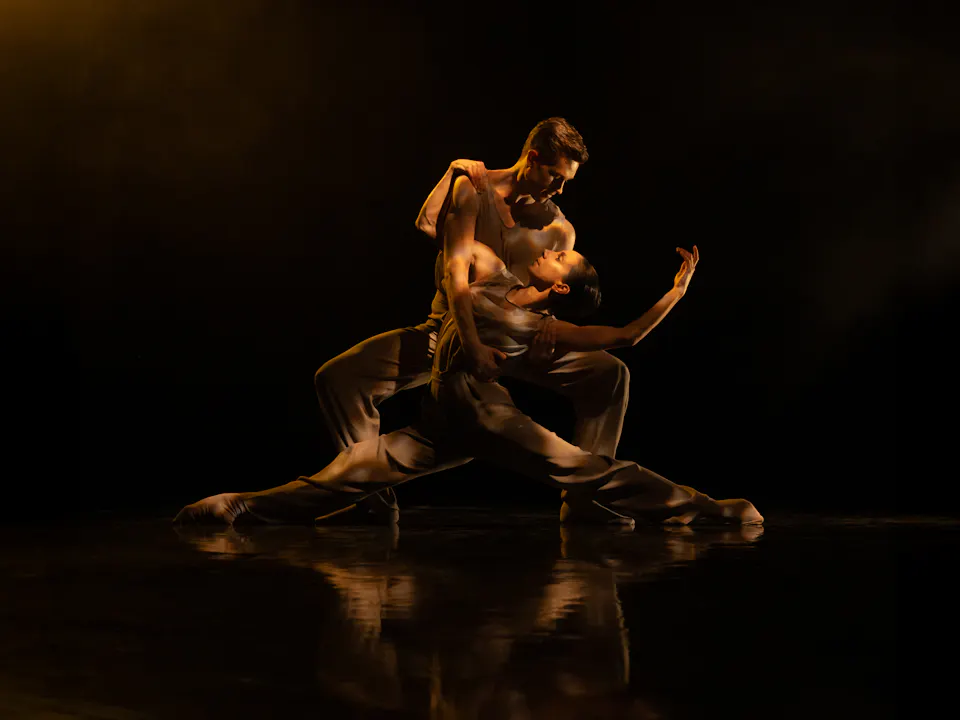 Two ballet dancers embrace passionately on a dark stage. One dancer draped themselves over the other.