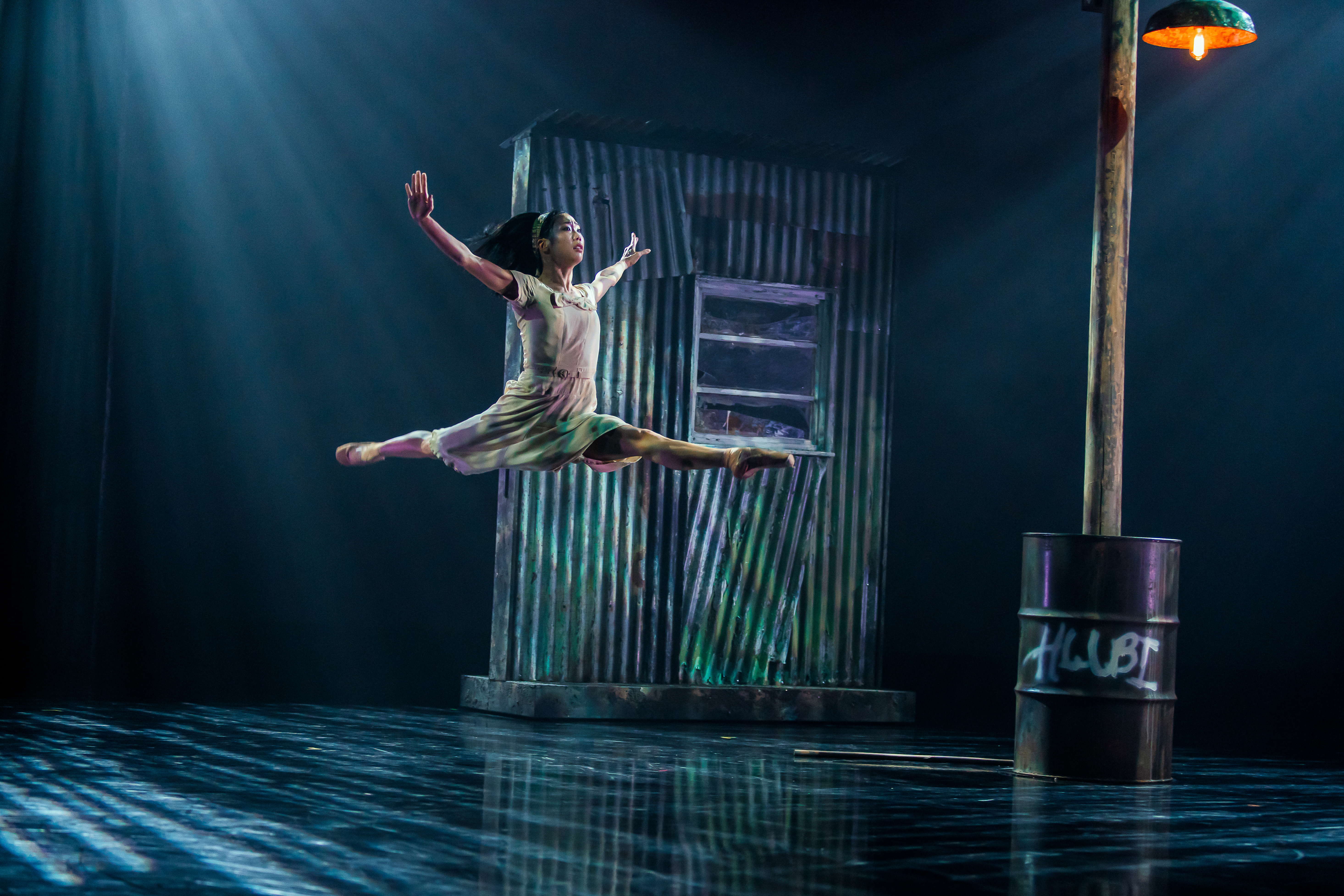 A ballet dancer launches themselves into the air on a dimly lit stage. Behind them on the stage is a corrugated iron shed and a rusted barrel holding up a wooden lamp post. (2 of 3)