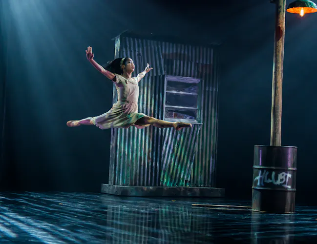 A ballet dancer launches themselves into the air on a dimly lit stage. Behind them on the stage is a corrugated iron shed and a rusted barrel holding up a wooden lamp post. (2 of 3)