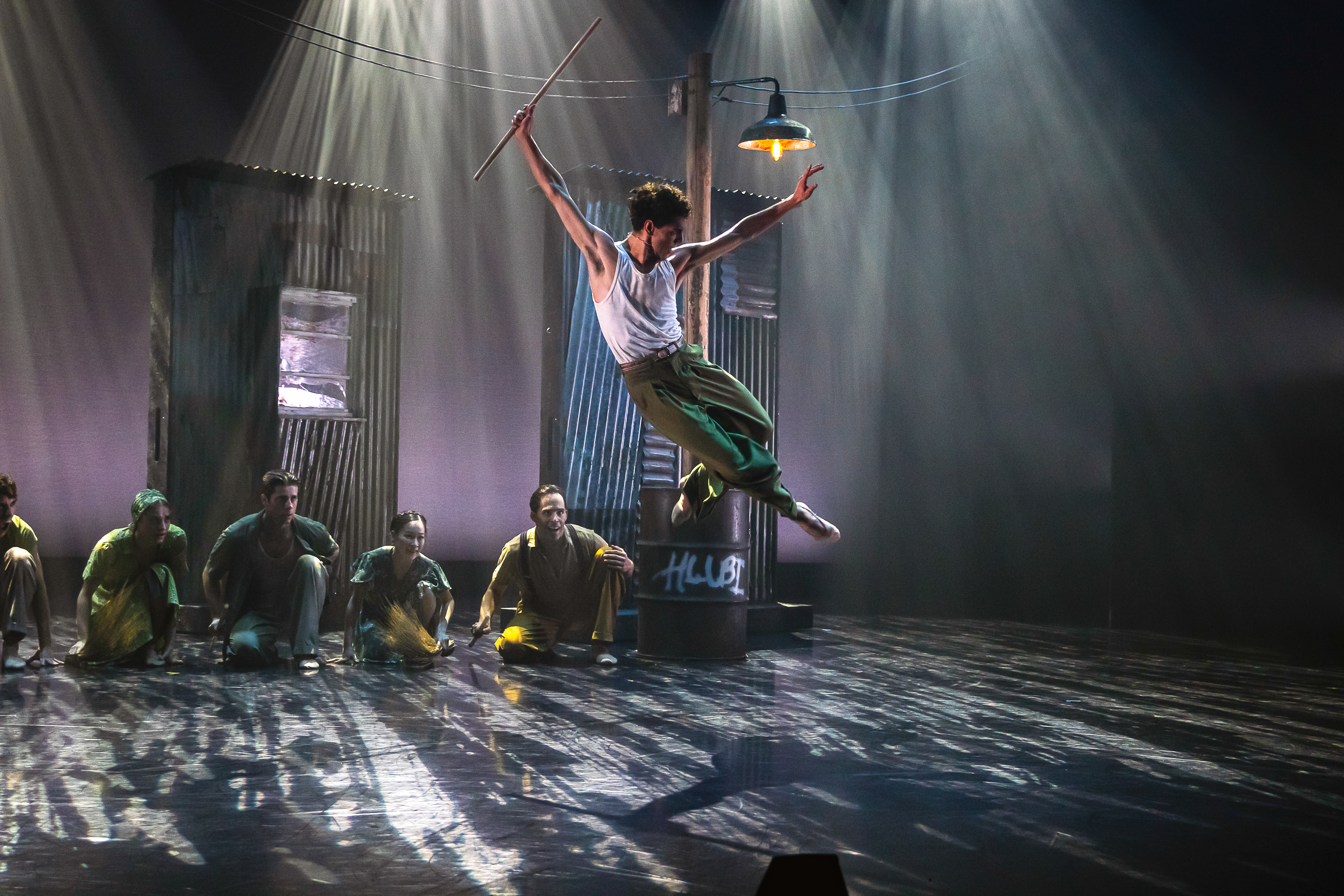 A ballet dancer launches themselves into the air on a dimly lit stage with haze. Several dancers sit on the floor watching. (3 of 3)