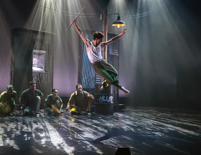 A ballet dancer launches themselves into the air on a dimly lit stage with haze. Several dancers sit on the floor watching. (3 of 3)