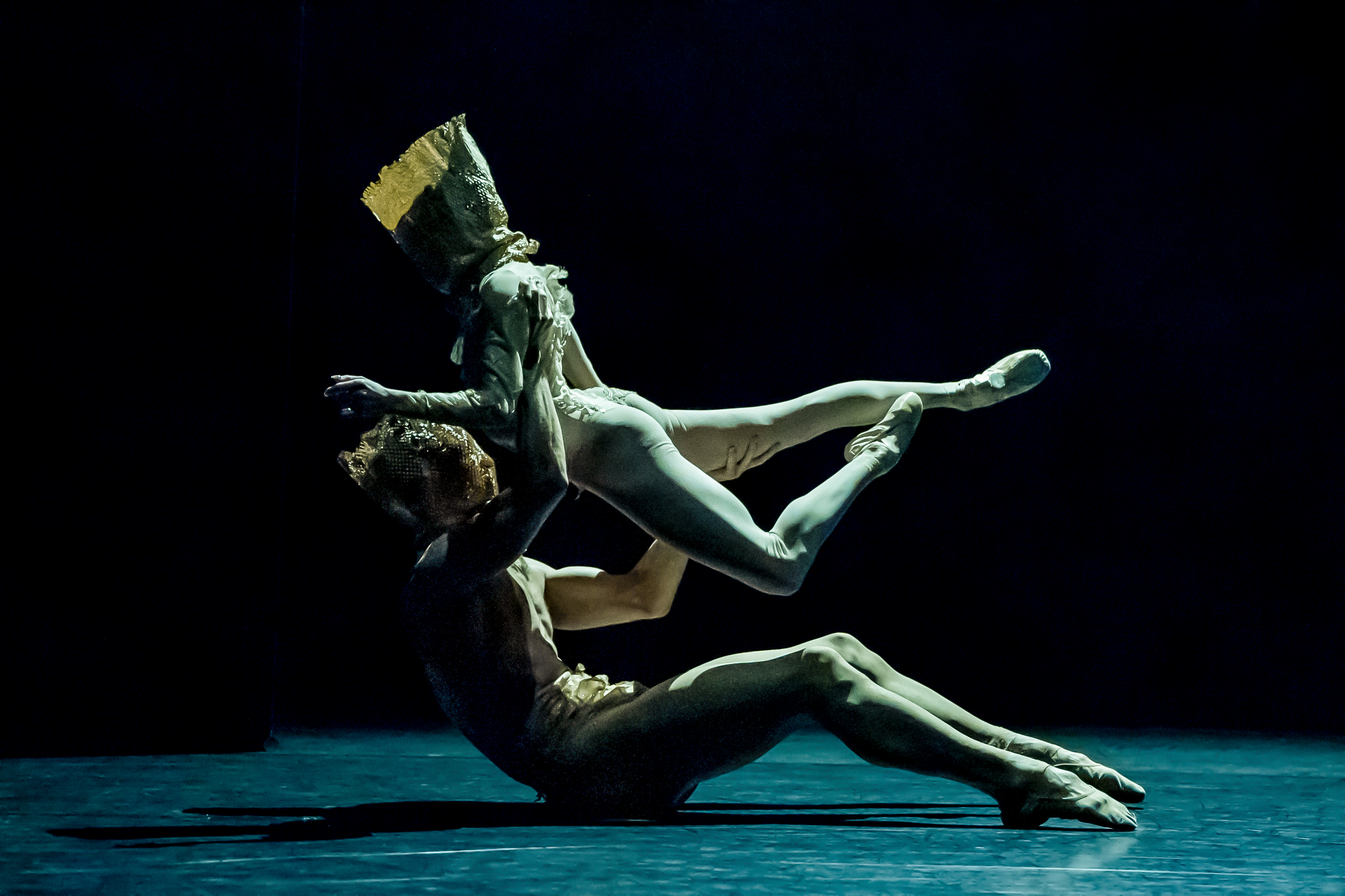 Two ballet dancers are on a dark stage wearing gold costumes. One lies on the floor lifting the other up with their legs stretched behind them.