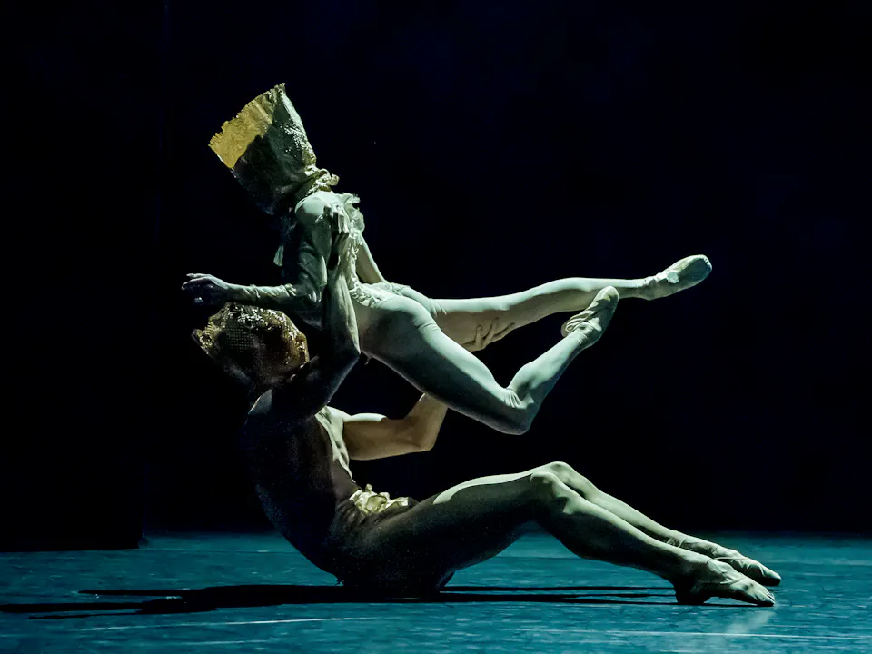 Two ballet dancers are on a dark stage wearing gold costumes. One lies on the floor lifting the other up with their legs stretched behind them.
