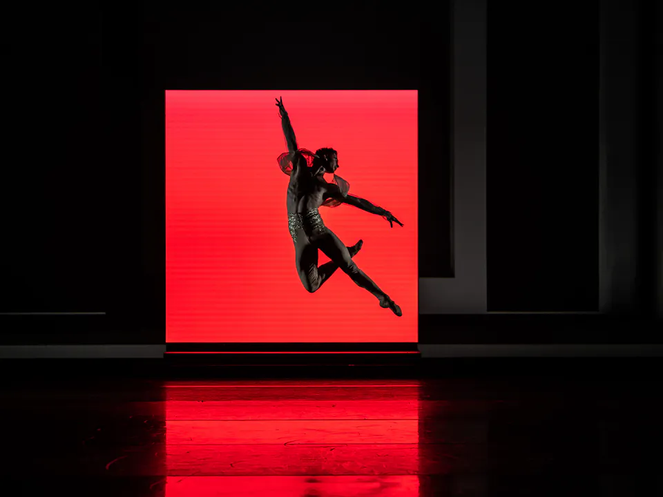 A ballet dancer launches themselves into the air on a dark stage in front of a square shape that is brightly lit red.