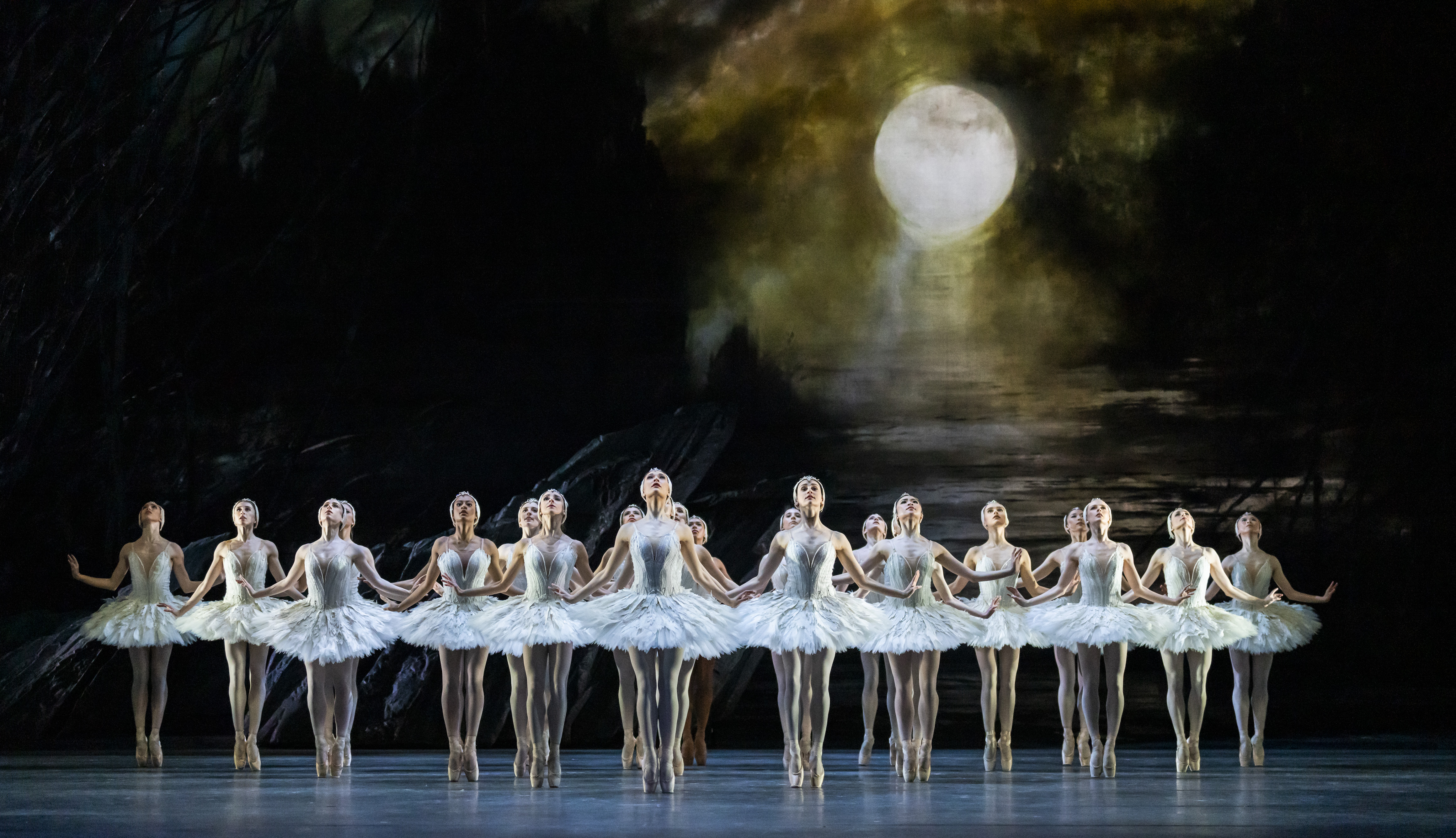 A dark set stage with several ballet dancers wearing white tutus and white feathered headdresses standing en pointe in lines. They perform in front of a dark rock front and the background is dark, moody skies. They are performing in The Royal Ballet's production of Swan Lake. (1 of 10)